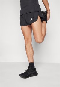 Black athletic shorts with a fitted inner lining and side slits. Worn with black socks and sneakers. Short, textured, and lightweight design.