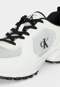Λευκό και γκρι αθλητικό παπούτσι με μαύρα κορδόνια και λογότυπο στο πλάι και στη γλώσσα, με σχεδιασμό υφασμένου διχτυού και παχύ μαύρο πέλμα.