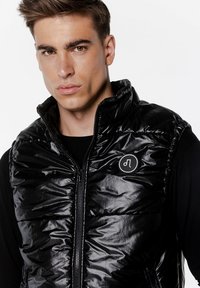 Czarna kamizelka puchowa o błyszczącej, pikowanej fakturze i wysokim kołnierzu. Posiada zamek błyskawiczny z przodu oraz okrągłe logo po lewej stronie.