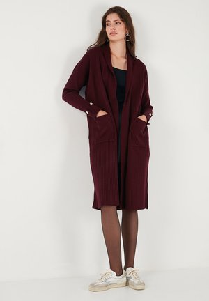 Burgundy ribbed cardigan with long sleeves, open front, two pockets, and button details on cuffs. Worn over a black dress and paired with sneakers.