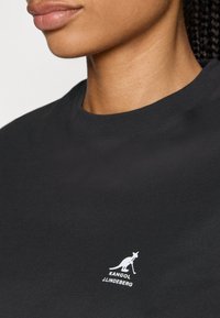 Juodas medvilnės marškinėlis su apvalia iškirpte, ant krūtinės yra mažas baltas kengūros logotipas ir užrašas "KANGOL J.LINDEBERG".