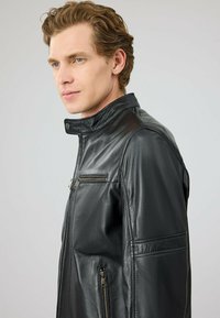 Zwart leren jack met hoge kraag, voorrits en borstzak, met een gladde textuur en gestikte details langs de mouwen.