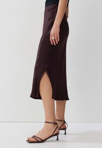 Brauner Satin-Midirock mit seitlichem Schlitz, glatte Textur, schwingender Saum, kombiniert mit braunen Riemchensandalen mit Absatz, Modell steht auf einer weißen Fläche.