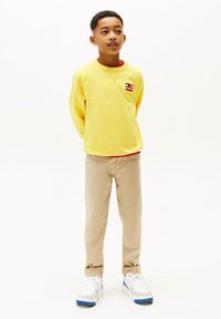 Sweatshirt jaune avec un petit logo brodé, chemise rouge en dessous, pantalons beiges et baskets blanches avec des accents bleus.