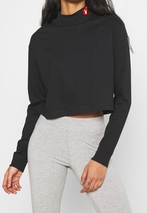 Vrouw draagt een zwarte cropped top met lange mouwen en een rood logo op de kraag, gecombineerd met lichtgrijze nauwsluitende broek.