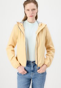 Giacca leggera gialla con cappuccio, fodera grigia, polsini elastici, cerniera completa, indossata sopra una camicia azzurro chiaro, abbinata a jeans blu.