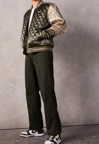 Vadderad bomberjacka i mörkgrön och beige med ribbade mudd, tillsammans med olivgrön byxor och vita/svarta sneakers.