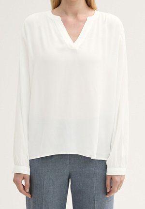 Biała bluzka z długim rękawem i dekoltem w serek, noszona z szarymi, dopasowanymi spodniami, pokazująca górną część ciała i ramiona na neutralnym tle.