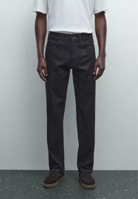 Black denim jeans with a straight leg fit, featuring front pockets and a button closure, paired with a light gray T-shirt and dark shoes.
