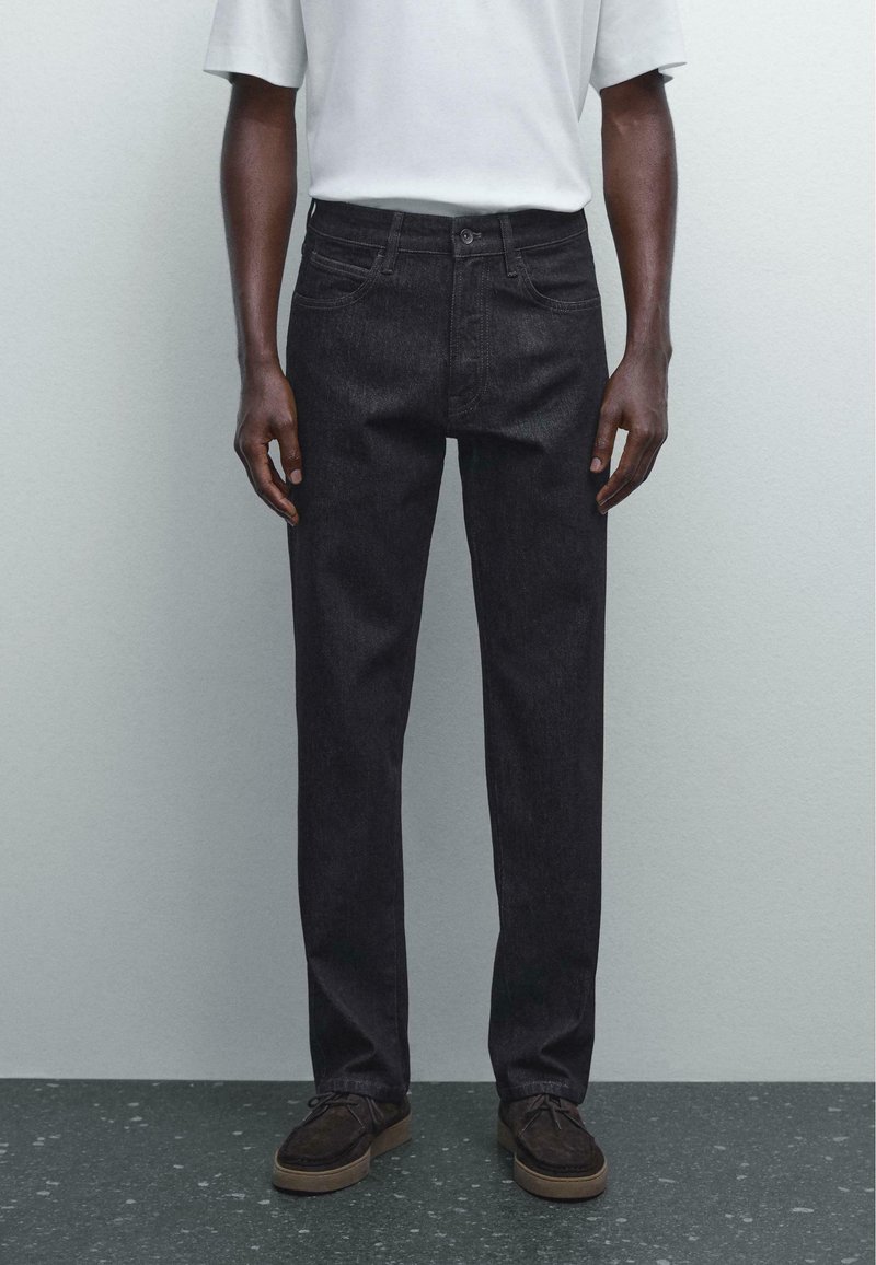 Black denim jeans with a straight leg fit, featuring front pockets and a button closure, paired with a light gray T-shirt and dark shoes.