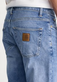 Pantalones vaqueros de mezclilla azul claro con un bolsillo trasero cosido que incluye un pequeño parche cuadrado de cuero marrón con el logo de Carhartt.