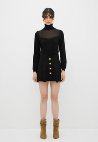 Haut noir à manches longues et col roulé avec un décolleté transparent, associé à une jupe noire taille haute ornée d'une rangée de boutons dorés. Bottes à talons marron.