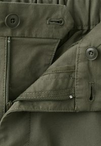 Gros plan sur un tissu vert montrant une fermeture éclair partiellement ouverte et deux boutons avec leurs boutonnières adjacentes sur un vêtement.