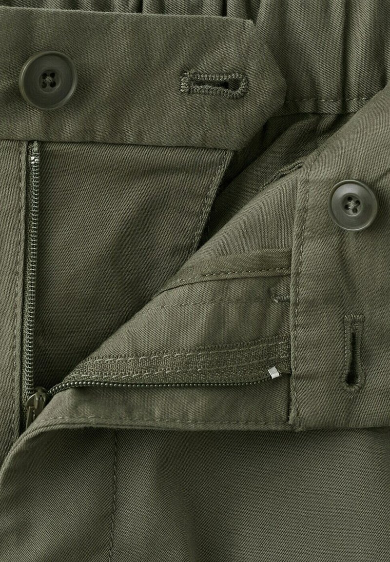 Gros plan sur un tissu vert montrant une fermeture éclair partiellement ouverte et deux boutons avec leurs boutonnières adjacentes sur un vêtement.