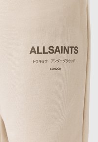 Beige stof med sort trykt tekst, der læser "ALLSAINTS," japanske tegn, og "LONDON" på tøj detalje.