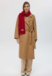 Cappotto in lana color cammello con vita cinturata, maniche lunghe e tasche laterali, abbinato a una sciarpa rossa con etichetta logo.
