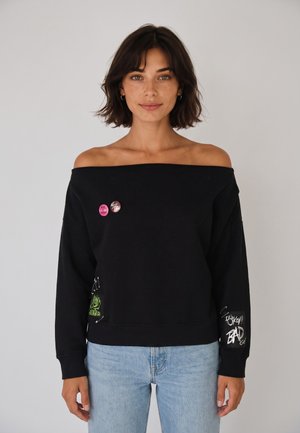 Donna con capelli scuri e corti che indossa un maglione nero scollato con spille e jeans azzurri, in piedi davanti a uno sfondo semplice.