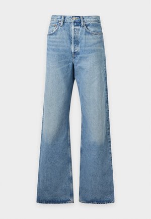 Donna con capelli lunghi e scuri che indossa una maglia grigio chiaro a maniche lunghe, jeans blu a vita alta e gamba larga, e scarpe nere con punta, in piedi con una mano in tasca.