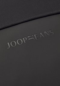 Črna tkanina z teksturirano površino in subtilnim črtastim vzorcem. Sestavljeno ime blagovne znamke "JOOP JEANS".