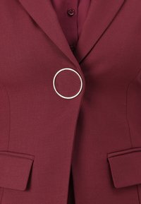 Blazer burdeos hecho de tela texturizada, con un adorno circular plateado en el cierre y dos bolsillos delanteros.