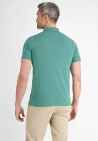 Zielona koszulka polo z krótkimi rękawami, gładką teksturą, płaskim kołnierzykiem i bocznymi rozcięciami, noszona z beżowymi spodniami. Widok z tyłu modela.