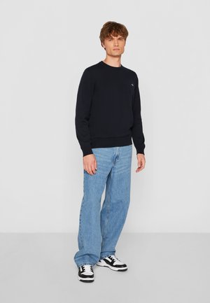 Marineblauer Strickpullover mit Rundhalsausschnitt, kombiniert mit hellblauen Jeans. Die Sneakers haben schwarze und weiße Akzente und ein flaches Sohlen-Design.