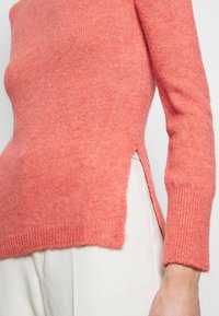 Maglione a maglia corallo con maniche lunghe, spacchi laterali e polsini a coste. Texture morbida e vestibilità leggermente larga. Senza cuciture sulle spalle.