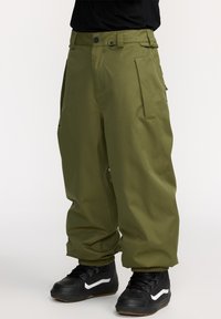 Olivgrüne Cargo-Hosen mit lockerer Passform, die mehrere Taschen, elastische Bündchen und einen Knopfverschluss aufweisen, getragen mit schwarzen Sneakern.
