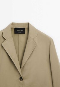 Jasnobeżowy blezer z klapami, zapinany na jeden guzik, o gładkiej fakturze. Wewnątrz widoczna metka z nazwą marki.