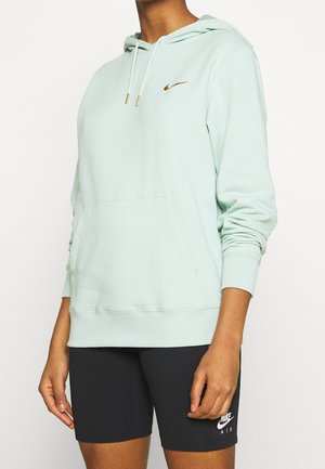 Haut à capuche vert clair avec une grande poche avant, capuche ajustable et un logo Nike doré sur la poitrine, associé à des shorts de vélo noirs à côtes.