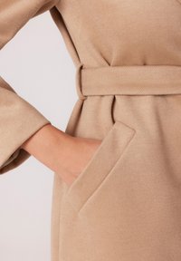 Beżowy płaszcz z wełny z paskiem w talii, bocznymi kieszeniami oraz miękkim, teksturowanym wykończeniem i luźnym krojem.