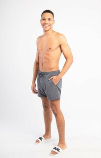 Grijze zwembroek met een elastische tailleband en zijzakken, gecombineerd met witte slippers met zwarte tekst. Model staat voor een neutrale achtergrond.