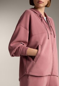 Ružová mikina s kapucňou a voľným strihom, zaobleným lemom, prednými vreckami a viditeľnými švami. Mäkko šitá z látky s matným povrchom.