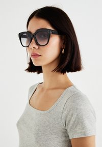 Óculos de sol oversized pretos com armação quadrada, lentes degradê e subtis detalhes em ouro, usados com uma blusa justa cinza claro.