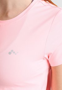 Różowa koszulka sportowa z okrągłym dekoltem, wykonana z gładkiego, lekkiego materiału. Posiada srebrne logo z małymi kropkami na lewej piersi.