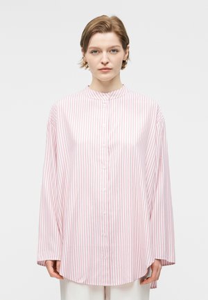 Femeie cu păr scurt purtând o cămașă largă cu dungi verticale roz și albe, cu nasturi, și pantaloni albi, stând pe un fundal simplu.