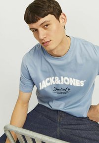 Camiseta de manga corta azul clara con el texto blanco "JACK & JONES" y "Studio Co." impreso en el frente. Lleva unos vaqueros oscuros.