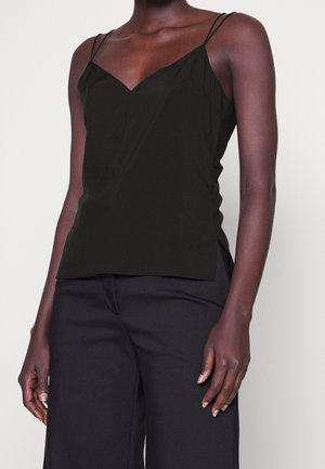 Dark-skinned person wearing a black sleeveless V-neck top with thin straps and black wide-leg pants, standing against a plain background.