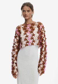 Crop top au crochet avec des manches longues, fond marron, motifs floraux superposés en rose et pêche, porté sur une robe blanche ajustée.