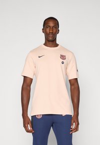 Mand iført lyserød FC Barcelona Nike T-shirt og blå bukser står overfor en ensfarvet lys baggrund.