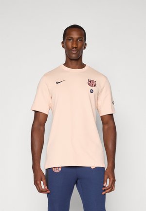 Homme portant un t-shirt Nike FC Barcelone rose clair et un pantalon bleu, se tenant devant un fond clair.