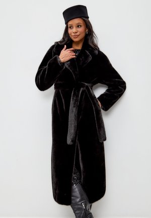 Cappotto in finta pelliccia nera con colletto a scialle, vita cinturata, tessuto testurizzato e spacco laterale. Indossato con un cappello nero abbinato e stivali neri.