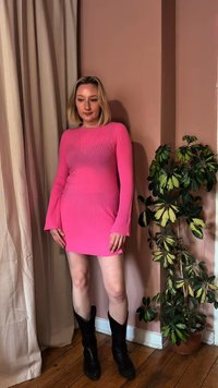 Γυναίκα που φοράει φωτεινό ροζ μίνι φόρεμα με μακριά μανίκια και μαύρες μπότες, στέκεται σε εσωτερικό χώρο δίπλα σε πολύχρωμο φυλλώδες φυτό πάνω σε ξύλινο πάτωμα.