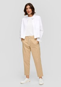 Witte overhemd met knoopjes over een gestreept beige en witte top, gecombineerd met beige broek met touwtjes en witte sneakers, tegen een grijze achtergrond.