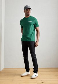 Green cotton t-shirt with a crew neck and white logo, paired with black jeans and white sneakers. Model wearing a gray cap.