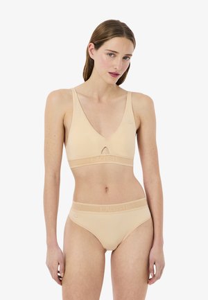 Mujer con conjunto beige de sujetador deportivo y bragas con bandas con logo, de pie sobre fondo blanco liso, mirando al frente con postura relajada.