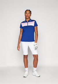 Polo shirt da golf blu con righe orizzontali rosa e bianche, maniche corte e colletto con zip. Indossato con pantaloni corti bianchi e guanto da golf.