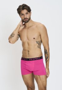 Shorts in cotone rosa con una fascia elastica nera che presenta la parola "RESPECT" in lettere rosa. Texture liscia e design aderente.