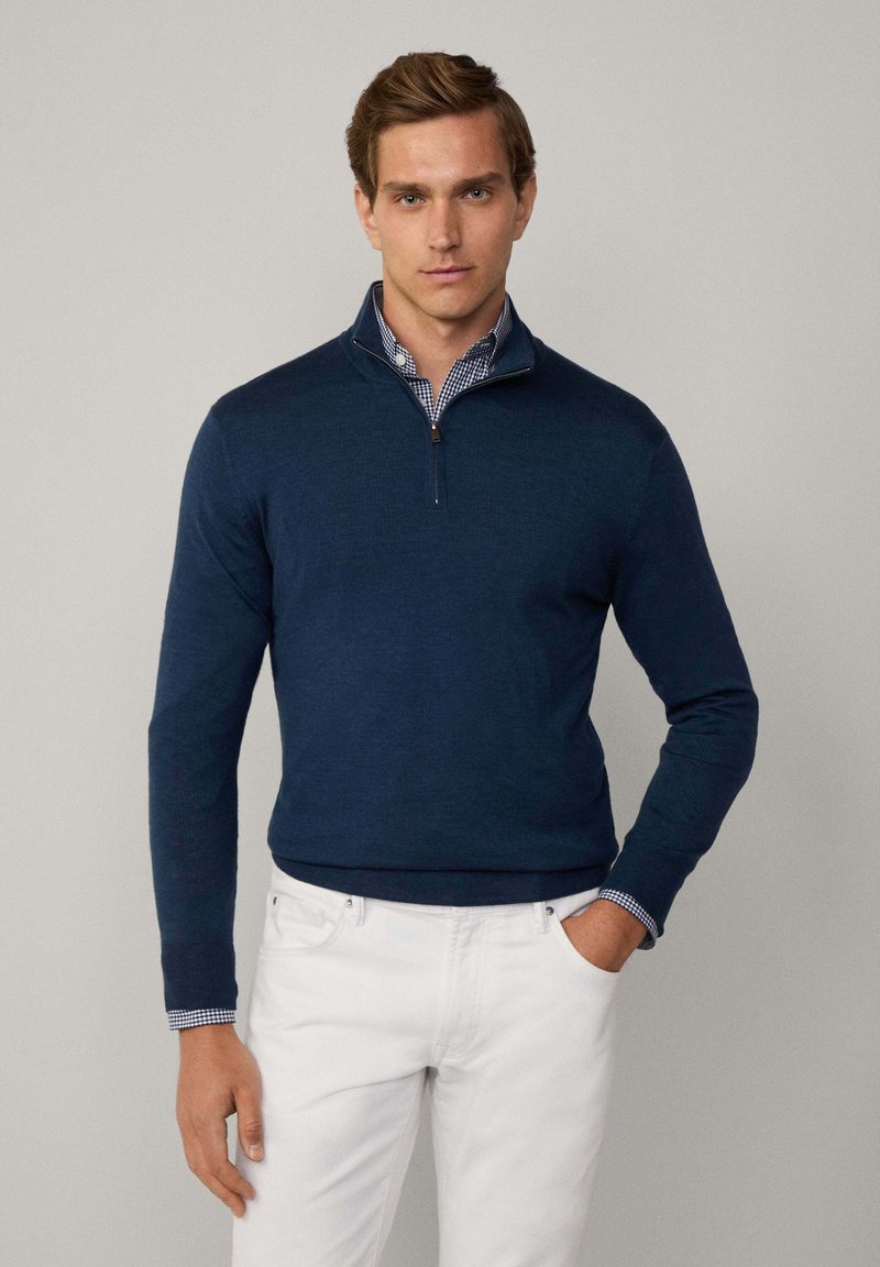 Marineblauer Pullover mit Reißverschlusskragen, aus einem strukturierten Stoff. Getragen über einem blau-weißen karierten Hemd, kombiniert mit weißen Hosen.