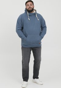 Sudadera azul con bolsillo canguro, cordones y un forro interior de contraste, combinada con vaqueros oscuros y zapatillas blancas.